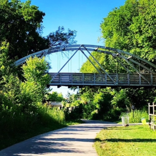 A metal arch bridge spans a tree-lined path, surrounded by lush greenery and blue skies.