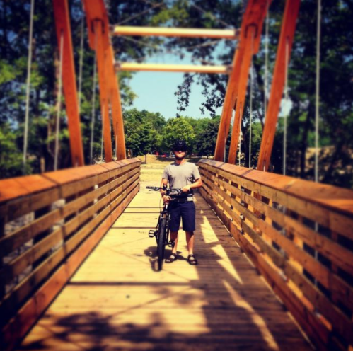 A person stands with a bicycle on a wooden bridge surrounded by trees on a sunny day.