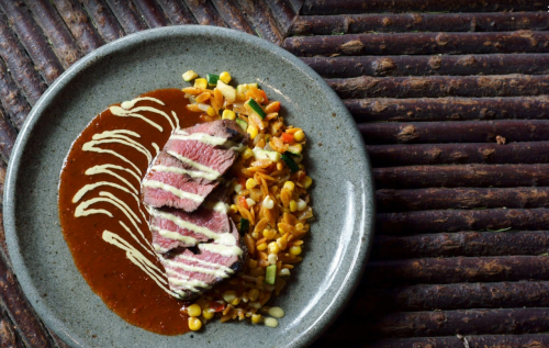A plate of sliced steak drizzled with sauce, served with a colorful grain and vegetable medley.