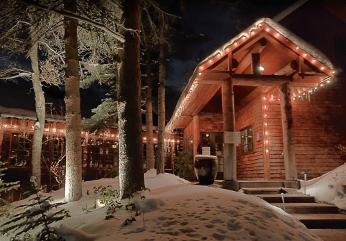 A cozy wooden cabin adorned with string lights, surrounded by snow-covered trees at night.