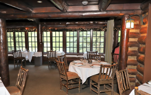 Cozy restaurant interior with wooden beams, tables set for dining, and large windows overlooking greenery.