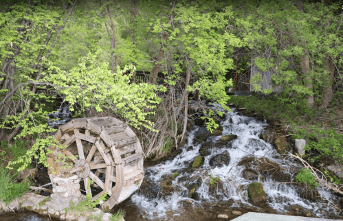 A wooden water wheel beside a flowing stream, surrounded by lush green trees and rocks.