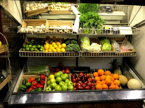 A display of fresh fruits and vegetables, including greens, apples, oranges, and peppers, in a market setting.