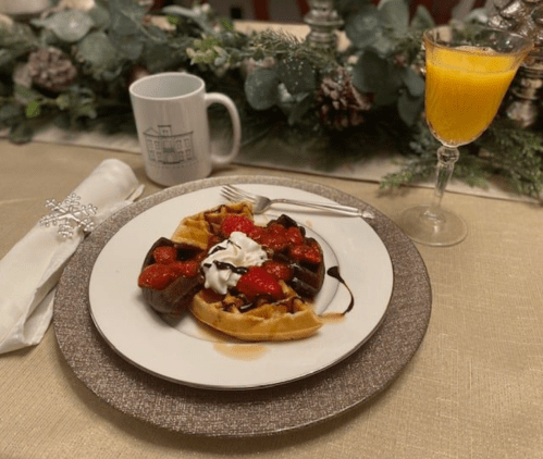 A plate with waffles topped with strawberries, whipped cream, and chocolate sauce, alongside a glass of orange juice.