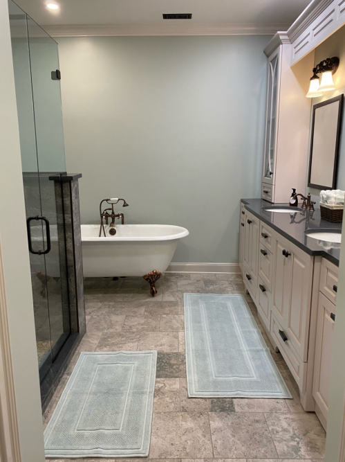 A modern bathroom featuring a freestanding bathtub, glass shower, and light-colored cabinetry with soft blue rugs.