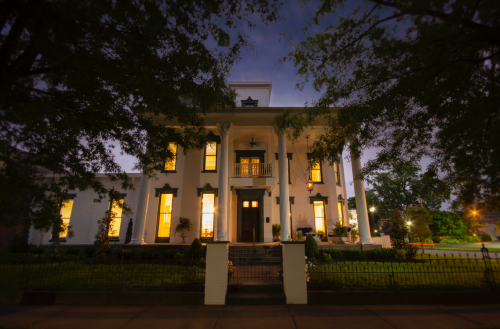 A grand white mansion with columns, illuminated windows, surrounded by trees at dusk.