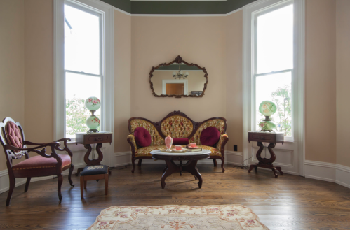 A vintage living room with ornate furniture, a round table, and large windows letting in natural light.