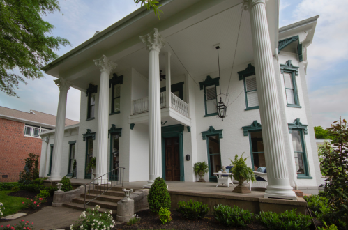 A grand white house with tall columns, green accents, and a welcoming porch surrounded by lush greenery and flowers.