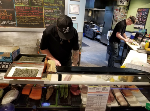 Two chefs prepare sushi in a restaurant kitchen, with fresh ingredients displayed in a glass case.