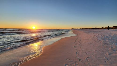 A serene beach at sunset, with gentle waves and footprints in the sand under a colorful sky.
