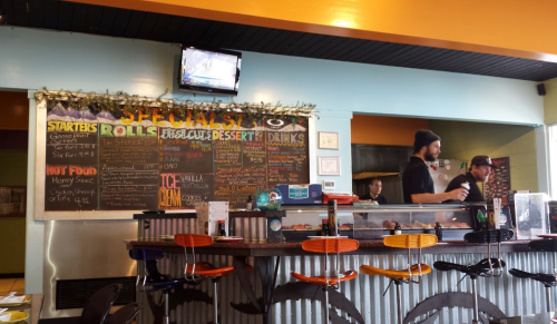 A colorful restaurant interior featuring a menu board, bar seating, and staff preparing food. A TV is mounted on the wall.