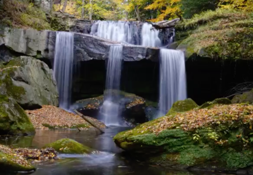 A serene waterfall cascades over rocks, surrounded by autumn foliage and a calm pool of water below.