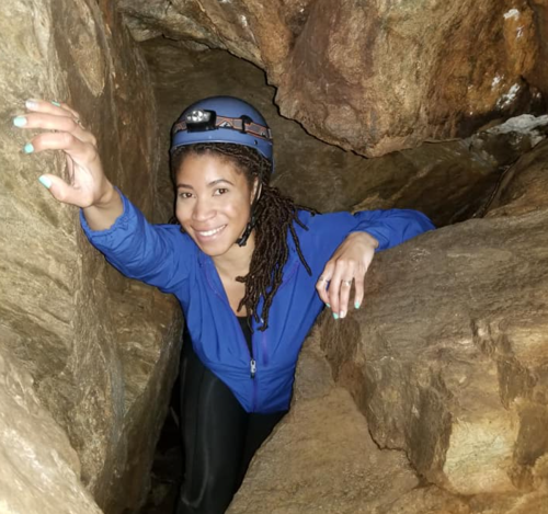 A person in a blue jacket and helmet climbs through a narrow cave passage, surrounded by rocky walls.