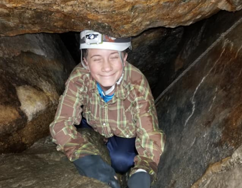A person in a helmet smiles while crawling through a narrow cave passage, surrounded by rocky walls.