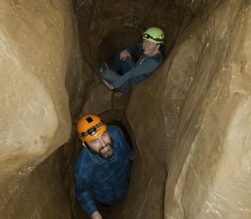 Two people in helmets exploring a narrow cave passage, with one looking up at the camera and the other seated above.
