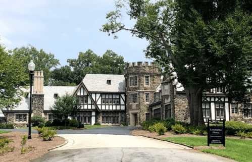 A large, historic stone building with a tower, surrounded by trees and a well-maintained landscape.