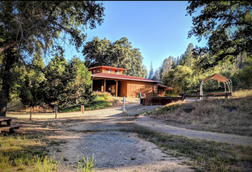 A rustic wooden building surrounded by trees, with a gravel path leading up to it and a picnic area nearby.