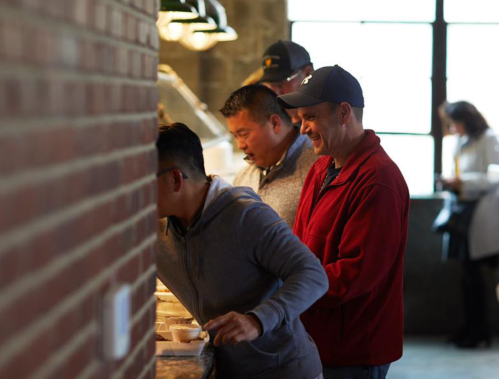 Three men serve themselves at a buffet, with a brick wall in the foreground and a window in the background.