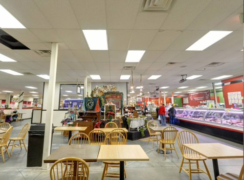 A spacious market interior with tables, chairs, and a meat counter in the background, featuring shoppers and bright lighting.