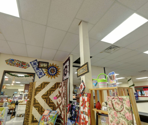 Interior of a store featuring quilts, crafts, and decorative items, with a ceiling and a welcoming sign.