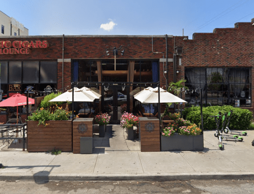 A cozy restaurant with outdoor seating, colorful flowers, and umbrellas, located on a city street.