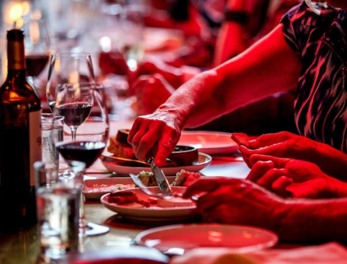 A close-up of hands at a dinner table, with plates of food, wine glasses, and a warm, red ambiance.