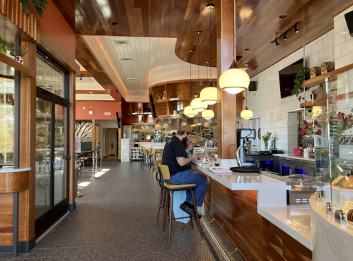 A cozy café interior with wooden accents, featuring a man sitting at the counter and a well-lit dining area in the background.