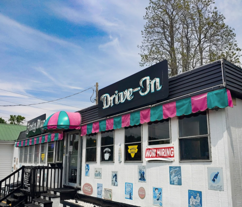 A colorful drive-in restaurant with a sign, "Drive-In," and a hiring notice, set against a clear blue sky.