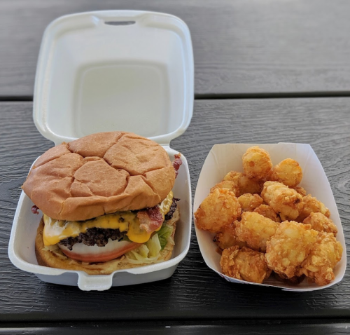 A takeout container with a cheeseburger and a side of crispy tater tots on a wooden table.