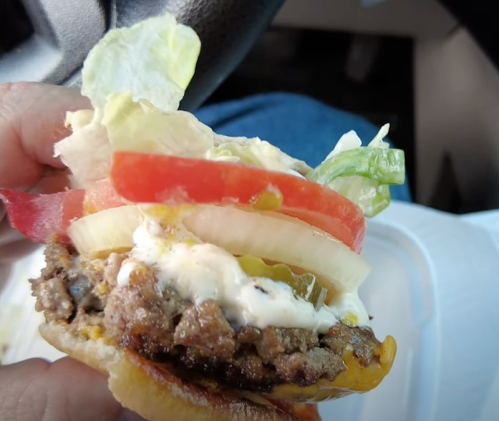 A close-up of a hand holding a burger with lettuce, tomato, onion, and sauce, set against a car interior background.