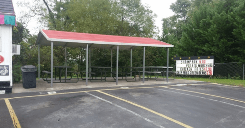 A covered picnic area with metal tables and benches, next to a sign advertising food and drinks. Trees in the background.