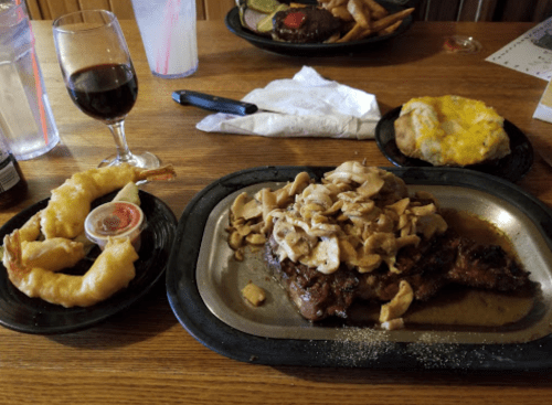 A plate with a steak topped with mushrooms, a baked potato with cheese, and a side of shrimp with dipping sauce.