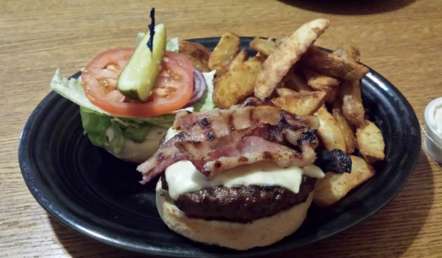 A plate with a bacon-topped burger, lettuce, tomato, pickles, and a side of crispy potato wedges.
