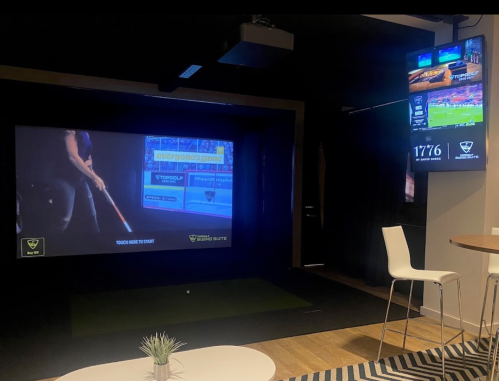 A modern indoor golf simulator setup with a large screen, a putting green, and a TV displaying sports information.