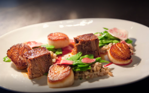 A gourmet dish featuring seared scallops, tender beef, greens, and radish on a white plate.