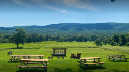 A scenic outdoor area with picnic tables, rocking chairs, and a backdrop of rolling green hills under a clear blue sky.