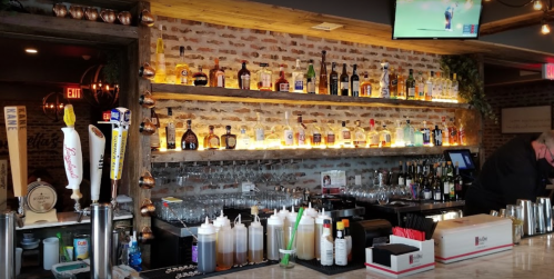 A well-stocked bar with various bottles on shelves, a TV screen, and a bartender preparing drinks.