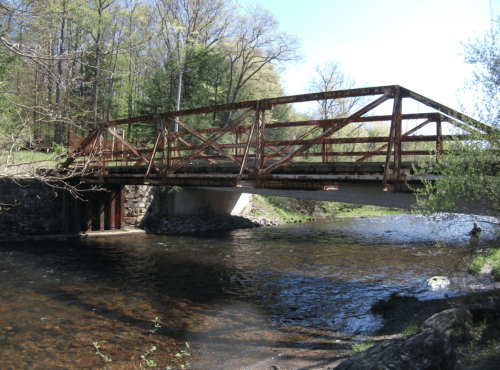 A rusty metal bridge spans a calm river, surrounded by lush greenery and trees under a clear blue sky.
