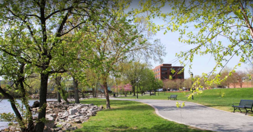 A scenic park with green trees, a pathway, benches, and a river, featuring a building in the background on a sunny day.