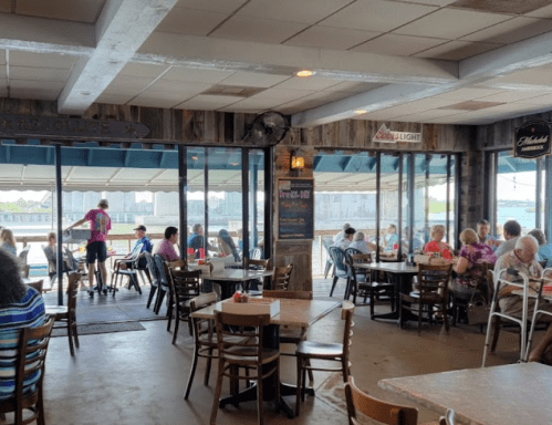 A bustling restaurant interior with diners at tables, large windows overlooking water, and a casual atmosphere.