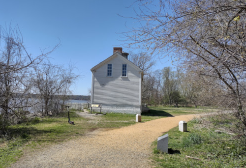 A historic white house beside a river, surrounded by bare trees and a gravel path on a clear day.