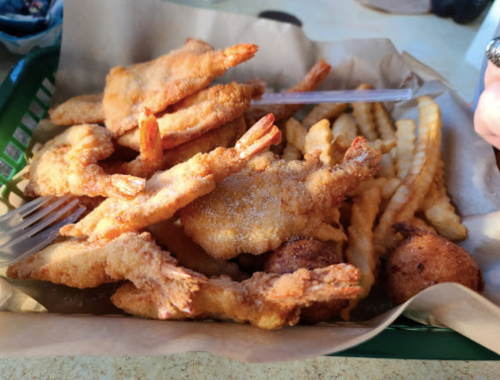 A basket of fried shrimp, hushpuppies, and crinkle-cut fries on brown paper.