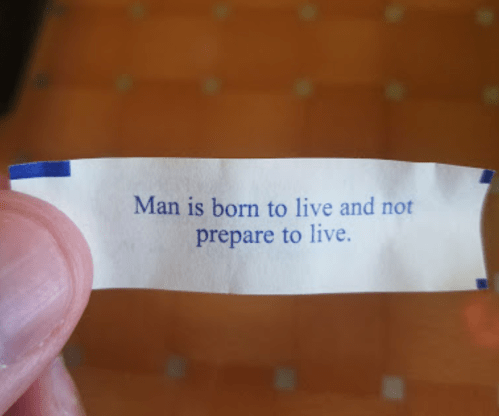 A hand holding a fortune cookie slip that reads, "Man is born to live and not prepare to live."