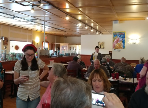 A busy restaurant scene with diners at tables, a woman standing with a phone, and a waiter in the background.