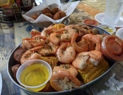 A large platter of shrimp, corn, sausage, and dipping sauces, served with hushpuppies in the background.