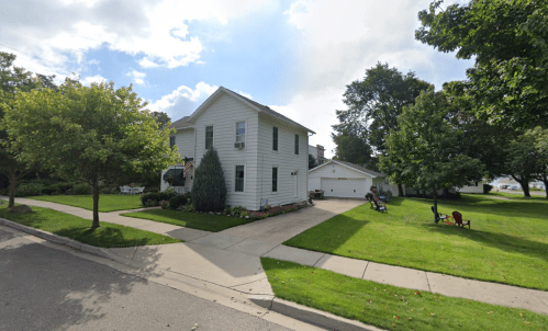A two-story white house with a green lawn, surrounded by trees, and a garage in a quiet neighborhood.