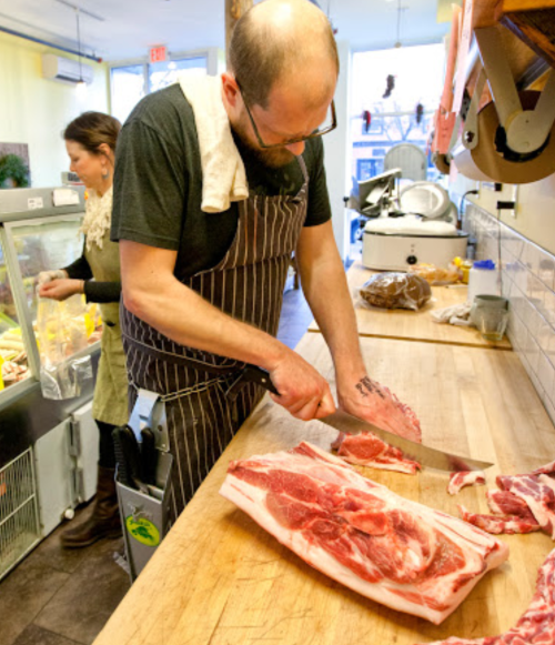 A butcher in an apron slices meat on a wooden counter, while a woman prepares food in the background.