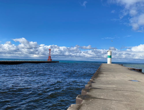 A view of a pier extending into the water, with a lighthouse in the distance under a blue sky with clouds.