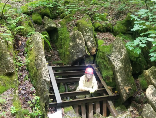 A person in a hard hat stands on a metal grate surrounded by moss-covered rocks in a forested area.