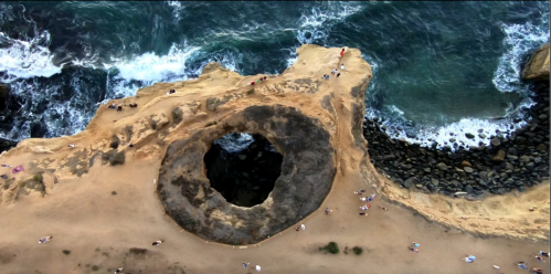 Aerial view of a rocky coastline with a large circular hole and waves crashing against the shore. Seagulls scattered around.